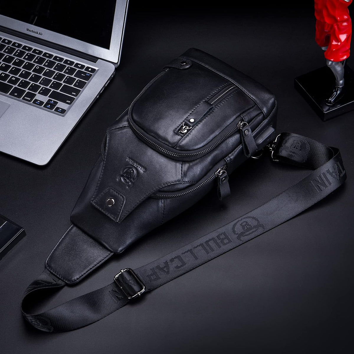 Black genuine leather sling bag on a desk next to a laptop, showing the front pockets and crossbody strap